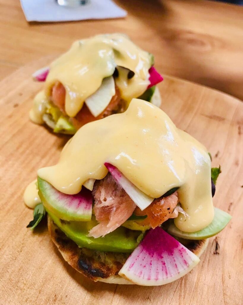 A burger with salmon, cheese, avocado and an interesting pink and green fruit or vegetable.